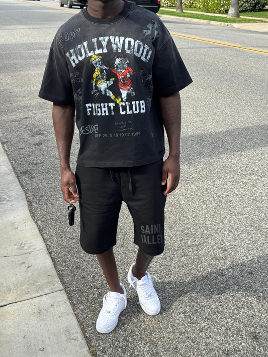Person wearing a 'Hollywood Fight Club' t-shirt and black shorts on a street.